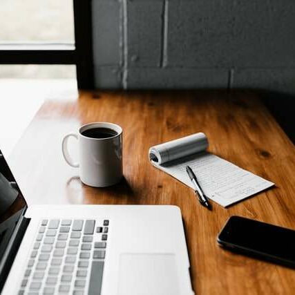 A laptop, notepad, pen, phone and a cup of coffee placed on a work desk